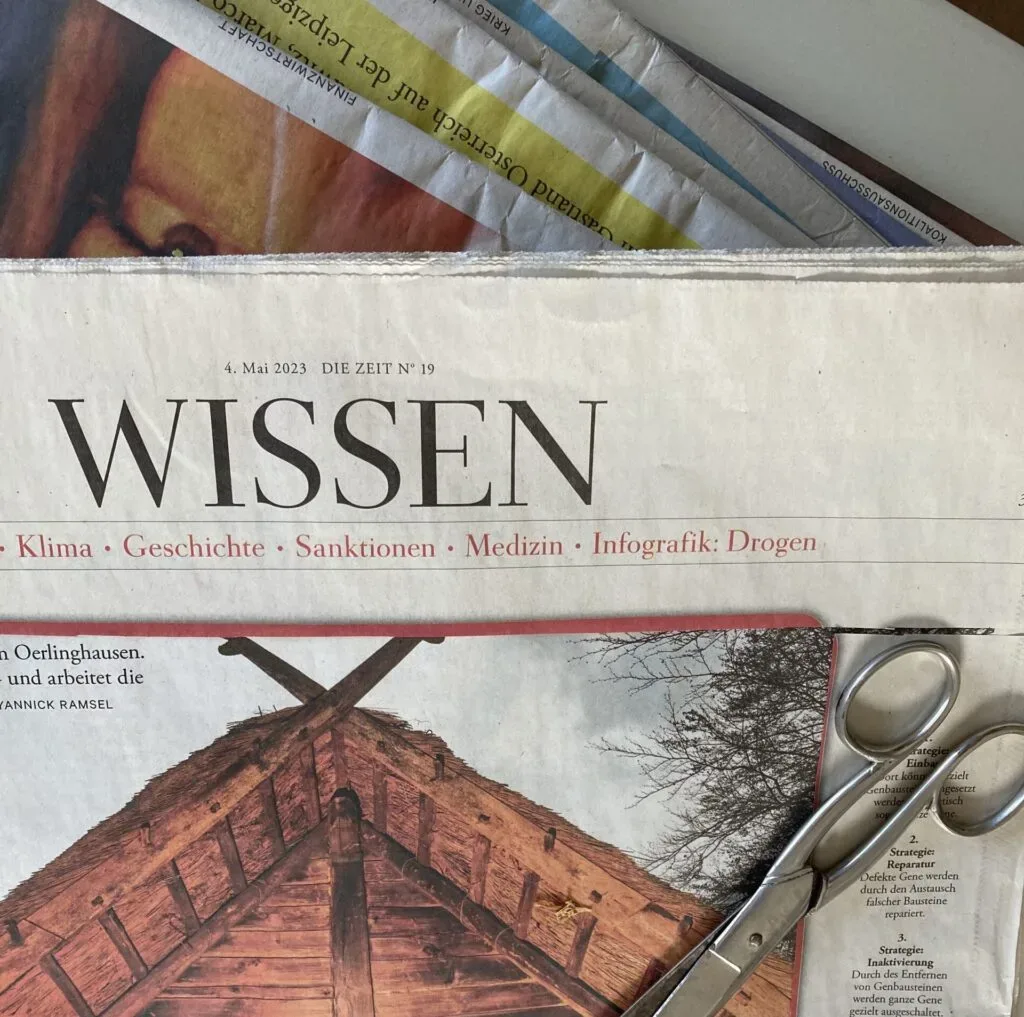 Zeitung mit Thema "Wissen" und Schere auf Tisch, teilweise verdeckt von Zeitschriften.
